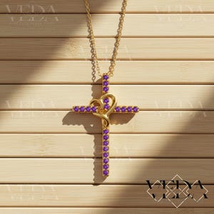 Pode incluir: Um colar com pingente dourado com um design de cruz e um detalhe em forma de coração. A cruz é adornada com pedras preciosas roxas. O colar pende de uma corrente dourada, sobre um fundo de madeira castanha clara.
