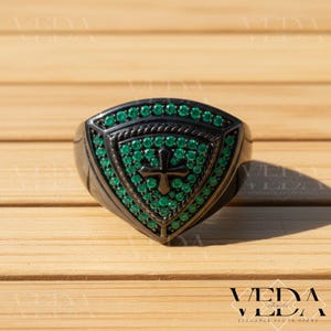 May include: A black ring with a shield-shaped face, decorated with green gemstones and a cross motif. The ring has a rope-like border and is set against a wooden surface. The ring is from Veda Jewels.