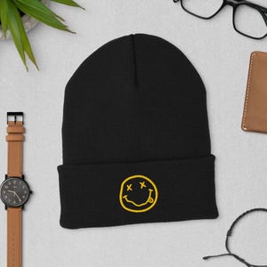 May include: Black beanie with a yellow embroidered smiley face design. The beanie is made of a soft, knitted material. The smiley face has two X's for eyes and a tongue sticking out. The beanie is a casual accessory.