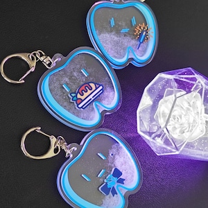 May include: Four acrylic keychains in the shape of an apple, each with a different food-themed design. The keychains have a blue and black border and silver hardware. A decorative, light-up, geometric rose is also visible.