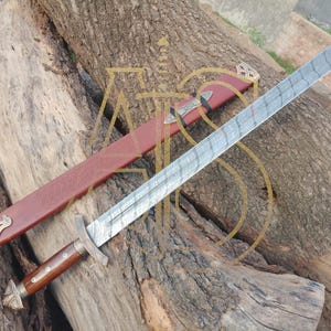 Pode incluir: Uma espada longa e prateada com uma lâmina estampada e um cabo de madeira, repousando sobre uma superfície de madeira. A espada tem uma guarda em forma de cruz e um pomo decorativo. Uma bainha de couro vermelha com detalhes dourados está ao lado.