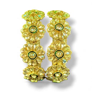 Puede incluir: Dos pulseras doradas con un diseño floral. Cada pulsera presenta múltiples adornos en forma de flor con piedras claras y facetadas y un centro verde. Las pulseras se muestran sobre un fondo blanco.