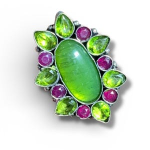 Peut inclure: Une bague en argent ornée d'une grande pierre verte ovale au centre, entourée de petites pierres vertes en forme de poire et de pierres roses foncées rondes. La monture de la bague est détaillée.