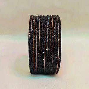 Puede incluir: Un conjunto de pulseras negras con una superficie brillante y texturizada. Las pulseras tienen un aspecto oscuro, casi iridiscente, con un sutil brillo metálico. El diseño presenta una serie de líneas paralelas, creando un efecto de capas.
