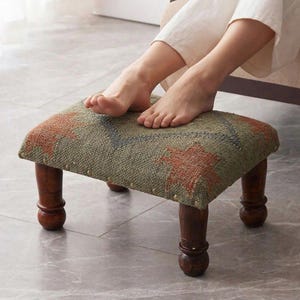 May include: A small, square footstool with a patterned fabric top in shades of green, orange, and blue. The footstool has four dark wooden legs. The person's feet are resting on the footstool.