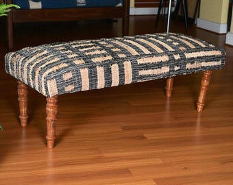Puede incluir: Un banco rectangular con una parte superior estampada y patas de madera. La parte superior de tela presenta un diseño geométrico en negro y beige. Las patas están torneadas y teñidas de color marrón cálido. El banco está sobre un suelo de parquet.