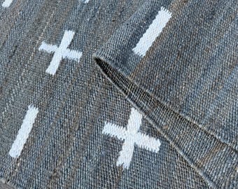 May include: A gray and white woven rug with a repeating pattern of white crosses and vertical lines. The rug is folded over, revealing the underside and the texture of the weave. The rug appears to be made of natural fibers.