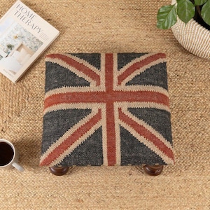 May include: A square footstool with a Union Jack design in gray, red, and beige. The footstool has four wooden legs and sits on a woven jute rug. A cup of coffee and a magazine are also on the rug.