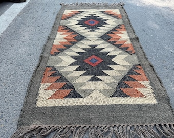 May include: Rectangular woven rug with a geometric pattern in shades of grey, black, orange, and cream. The rug features a central diamond design with fringed edges. The rug's dimensions are approximately 60 x 120 cm.