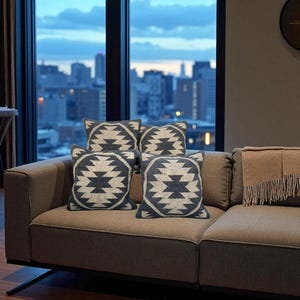 May include: Four square throw pillows with a geometric design in shades of gray and white are arranged on a brown sofa. A matching throw blanket with fringe is on the right side of the sofa. A cityscape is visible through the window.