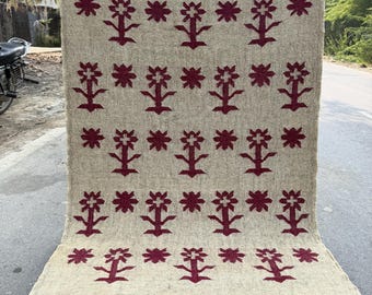 May include: A beige rug featuring a repeating pattern of maroon floral motifs. The rug has a fringed border and is shown outdoors on a paved surface. The floral design is symmetrical and covers most of the rug's surface.