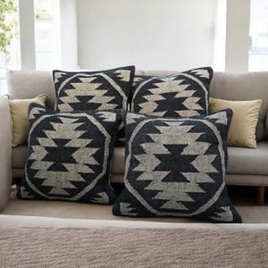 May include: Four square throw pillows with a geometric design in shades of charcoal gray and beige. The pillows are arranged on a light gray sofa, creating a cozy and stylish living room setting. The pillows add a touch of texture and pattern.
