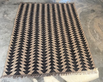 May include: Rectangular area rug with a repeating black and tan geometric pattern. The design features rows of inverted triangles. The rug has a fringed edge and is placed on a concrete surface.
