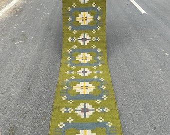 Könnte beinhalten: Ein langer, olivgrüner Läufer mit einem geometrischen Muster. Das Design zeigt sich wiederholende Quadrate in Blau, Weiß und Gelb. Der Teppich hat einen Fransenrand und wird im Freien auf einer Straße präsentiert.