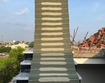 May include: A long, rectangular rug with a repeating pattern of light beige horizontal shapes on a dark green background. The rug is displayed outdoors, draped over steps, with a fringe at the bottom. The sky is blue.