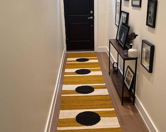 May include: A mustard yellow and white striped runner rug with large black circular accents. The rug is placed in a hallway with a black door and a dark wood console table with framed artwork.