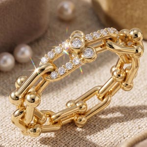 May include: A gold-toned ring featuring a chain-link design with spherical accents at each joint. The top section of the ring is embellished with a row of small, clear, sparkling stones set into the metal links, creating a bright, reflective surface.