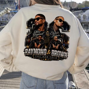 Back view of a person wearing a beige hoodie that displays the high-resolution Usher and Chris Brown 2026 concert tour design with sharp, contrasting metallic gold details.