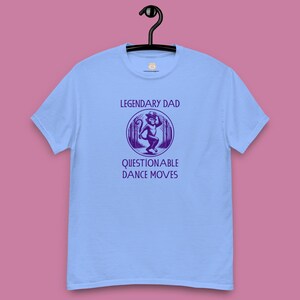 May include: Pale blue t-shirt featuring the text "LEGENDARY DAD" above a purple graphic of a monkey in a hat, and the words "QUESTIONABLE DANCE MOVES" below. The shirt is suspended from a black hanger.
