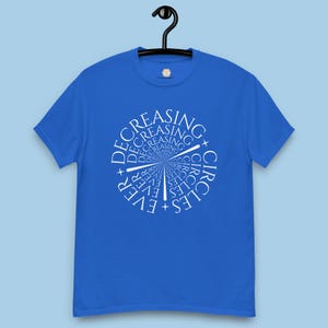 May include: A royal blue t-shirt with a white graphic design. The design features the words "EVER DECREASING CIRCLES" arranged in concentric circles and radiating lines. The shirt is hanging on a black hanger.