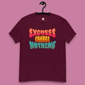May include: A maroon t-shirt with the text "EXCUSES CHANGES NOTHING" in orange, yellow, and teal lettering. The shirt is hanging on a black hanger against a pink background.