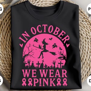 Może przedstawiać: Czarna koszulka z różowym motywem Halloween. Projekt przedstawia wiedźmę na miotle, nietoperze i napis "In October We Wear Pink" z różowymi wstążkami. Koszulka jest prezentowana na tkanej powierzchni.