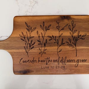 May include: Wooden cutting board with a handle, featuring engraved botanical illustrations and the text "Consider how the wildflower grow" from Luke 12:27-28. The board is rectangular with rounded corners.
