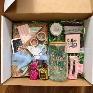May include: A gift box filled with various items, including a glass tumbler with the text "Not Perfect Just Forgiven", a pink hair clip, and a small notebook with the words "Follow the Shepard". The box also contains decorative paper and a blue ribbon.