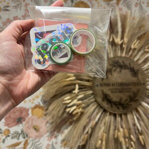 May include: A clear plastic bag containing stickers, washi tape, and a metal cross-shaped paper clip. The stickers have the word "Jesus" and other text. The washi tape is green and white. The background is a floral wallpaper.