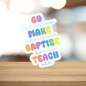 May include: A colorful sticker with the words "GO, MAKE, BAPTIZE, TEACH all" in a rainbow of colors. The sticker is on a wooden surface with a blurred background.