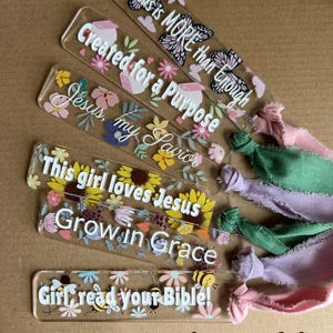 May include: Clear acrylic bookmarks with inspirational quotes and floral designs. Each bookmark features a different phrase, including "Jesus is MORE than Enough" and "Girl, read your Bible!" with attached colorful ribbons.