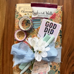 May include: A gift set wrapped in floral paper, tied with a light blue ribbon and a white flower. Includes washi tape, a card that says "GOD DID", and a sticker that says "CREATED WITH PURPOSE". Also includes a book and a bag of tea.