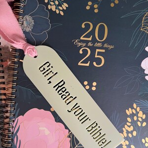 May include: A spiral-bound planner with a floral design in navy, pink, and gold. A light green bookmark with the text "Girl, Read your Bible!" is attached with a pink ribbon. The planner displays the numbers "2025".