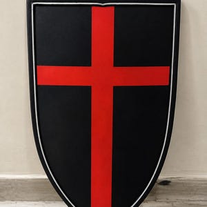 Pode incluir: Um escudo de estilo heater com uma superfície preta mate e uma cruz vermelha vertical e horizontal ousada centrada na parte frontal. O escudo é delineado com uma borda fina e elevada de cor prateada que segue a forma pontiaguda da base.