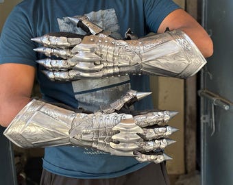 May include: A pair of silver-colored, articulated gauntlets with sharp, conical spikes on the knuckles and intricate engraved designs. The gauntlets are made of metal and appear to be part of a suit of armor, showcasing a medieval or fantasy theme.