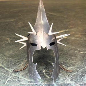 Puede incluir: Un casco de color plateado con una espiga central alta y múltiples espigas más pequeñas. El casco tiene aberturas para los ojos recortadas y un borde inferior dentado. El diseño sugiere un tema de fantasía o medieval.