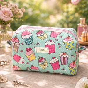 Könnte beinhalten: Eine rechteckige, mintgrüne Kosmetiktasche mit rosa Reißverschluss und Griffen. Die Tasche ist mit bunten Cupcakes und dem Text "1BIGN" gemustert. Der Hintergrund ist eine verschwommene Außenszene mit Blumen.