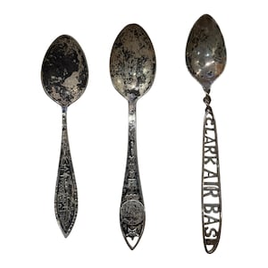 May include: Three silver-toned metal souvenir spoons with tarnished bowls. The left spoon features a building graphic, the centre spoon has a circular emblem and the text "MAH", and the right spoon has an openwork handle reading "CLARK AIR BASE".
