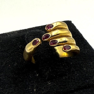 Puede incluir: Un anillo dorado con forma de mano, con piedras rojas en cada dedo. El anillo se exhibe sobre una superficie negra, resaltando su diseño detallado y el contraste de colores.