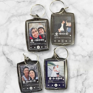 May include: Four clear acrylic keyrings with silver rings. Each keyring features a photo and a music player interface design. The photos show couples. The keyrings are on a white and grey marble surface.