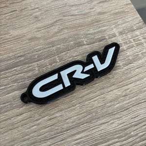 Pode incluir: Emblema CR-V em preto e branco. O emblema apresenta as letras "CR-V" em branco sobre um fundo preto. O emblema tem formato oval com um pequeno orifício numa extremidade.