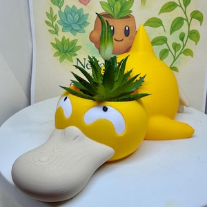 May include: A yellow, duck-like planter featuring a wide, beige bill and white eyes with black pupils. A green, spiky succulent plant sits inside the top opening of the head. The planter has a smooth, matte finish and a pointed tail-like structure.