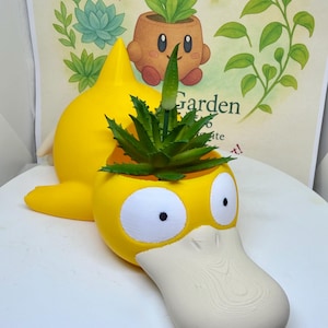 May include: A bright yellow planter shaped like a duck-billed creature with large white eyes and a beige bill. The planter features a hollow opening on the top containing a green succulent plant with a single thin stem extending upward from the center.