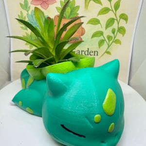 May include: A teal and lime green creature-shaped planter with a smooth finish, featuring a small artificial succulent plant nestled in the top opening. The planter has pointed ears, closed eyes, and light green spots on its head and back.