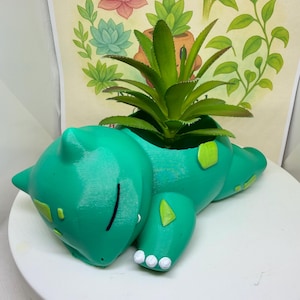 May include: A teal creature-shaped planter featuring a sleeping expression with closed eyes, small white claws, and light green patches on its back and limbs. A green succulent plant is potted inside the hollow opening on the creature's back.