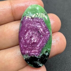 May include: An oval gemstone with a striking colour palette. The top is a vivid green, leading to a deep magenta centre with a swirling design. The base is black, providing a strong contrast. The gemstone is held in a hand.