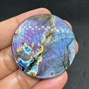 Puede incluir: Una piedra preciosa de labradorita redonda y pulida, con una fascinante exhibición de color. La piedra presenta una mezcla de azules, púrpuras y dorados, creando un efecto iridiscente y brillante. La superficie es lisa y reflectante, mostrando patrones naturales.