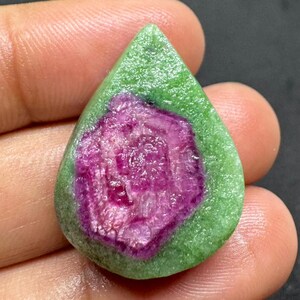 Puede incluir: Una gema en forma de lágrima con un vibrante centro magenta rodeado por un borde verde texturizado. La superficie de la piedra tiene un aspecto rugoso y natural, mostrando una mezcla de colores y texturas. La gema se sostiene en una mano.