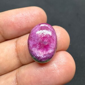 Könnte beinhalten: Ein oval geschliffener Edelstein mit einem Farbverlauf von Pink und Magenta. Die Oberfläche ist glatt und glänzend, mit feinen inneren Mustern. Der Edelstein wird in einer Hand vor dunklem Hintergrund gehalten.