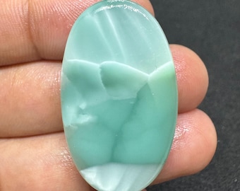 May include: Oval-shaped, polished gemstone with a light blue and white color pattern. The stone has a smooth, glossy surface and a unique, natural design. The gemstone is held in a hand, showcasing its size and shape. This gemstone is ideal for jewelry making.